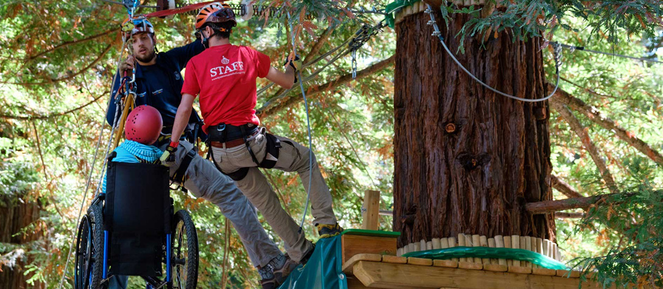 Due operatori del Dynamo Camp issano un bambino in carrozzina su un albero di un parco avventura - Enel.it